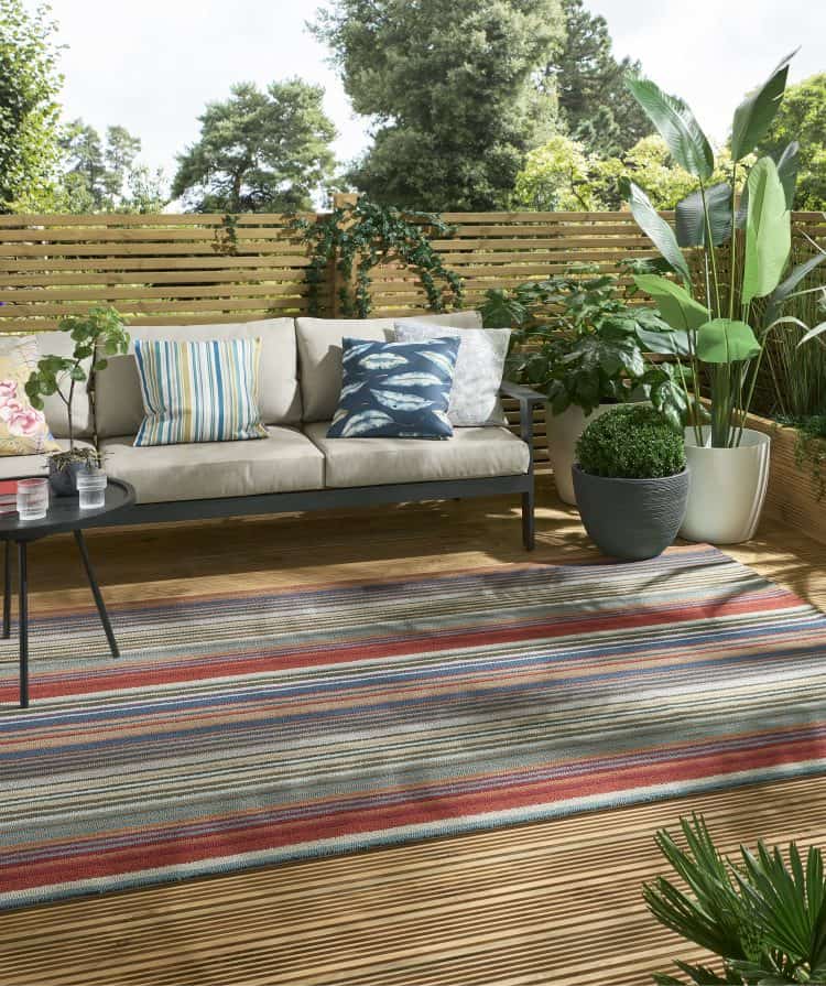 Teal and rust striped outdoor rug on wooden patio with plants and sofa.
